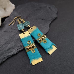 Peut inclure: Une paire de boucles d'oreilles pendantes composées de plaques rectangulaires en métal doré martelé enveloppées de tissu bleu sarcelle effiloché. Chaque boucle comporte trois petites perles dorées, des fils enroulés et une pierre bleue claire.