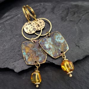 Könnte beinhalten: Ein Paar Ohrhänger mit goldfarbenen Klappbügeln, kleinen gehämmerten runden Scheiben und größeren quadratischen Platten mit einer verwitterten, oxidierten blau-bronzenen Patina. Jeder Ohrring ist mit einer einzelnen bernsteinfarbenen Perle versehen.