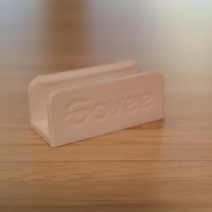 May include: A white, rectangular holder with rounded edges, featuring the embossed word "Govee". The holder is U-shaped, designed to hold a small object. It sits on a wooden surface.
