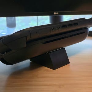 May include: A black handheld gaming device is mounted on a black charging dock. The device has a sleek, ergonomic design with visible buttons and ports. The setup is placed on a wooden surface, with a computer monitor in the background.