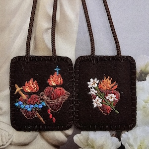 May include: Two dark brown embroidered religious pendants with brown cords. One features two hearts with flames, a cross, and a crown. The other has a heart with flames and white flowers. The embroidery is in red, orange, blue, and white.