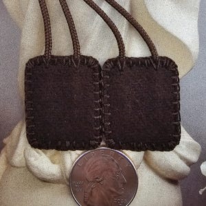 May include: Two dark brown, square scapulars with stitched edges and braided cords. A U.S. quarter coin is placed below the scapulars for size comparison. The scapulars are on a light background.