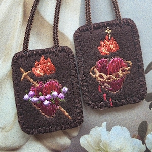 May include: Two rectangular brown felt scapulars featuring embroidered designs. One displays a heart pierced by a sword with purple flowers, and the other shows a heart encircled by thorns with a flame on top. Both have brown braided cords.