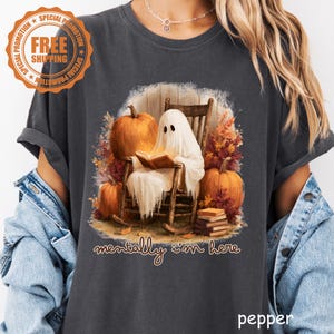 Op de afbeelding: Een antracietgrijs t-shirt met een afbeelding van een witte geest die in een houten schommelstoel zit en een boek leest, omringd door oranje pompoenen en herfstbladeren, met de tekst "mentally i'm here" eronder gedrukt.
