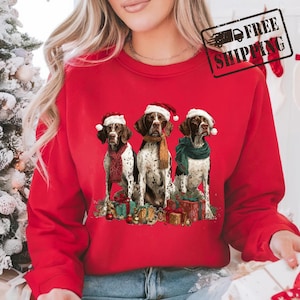May include: A red sweatshirt featuring three dogs wearing Santa hats and scarves, surrounded by wrapped gifts. The dogs are brown and white, with festive holiday attire. The image has a Christmas theme.