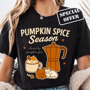 Puede incluir: Camiseta negra con un diseño gráfico que presenta el texto "PUMPKIN SPICE Season" y una cafetera, una taza de café con leche de calabaza y dos gatos. La camiseta también tiene el texto "Fueled by pumpkin spice."