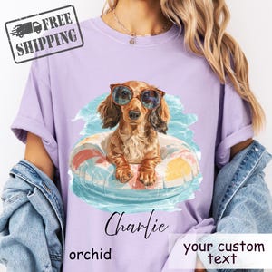 May include: Orchid-colored t-shirt featuring a dachshund wearing sunglasses and floating in a pool float. The shirt has the name "Charlie" and the words "your custom text" printed on it. The shirt is a comfortable fit.