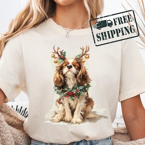 Può includere: T-shirt color crema con un Cavalier King Charles Spaniel che indossa una ghirlanda natalizia e corna con ornamenti dorati. Il cane ha gli occhi chiusi, suggerendo un'espressione pacifica. Il design è a tema festivo.