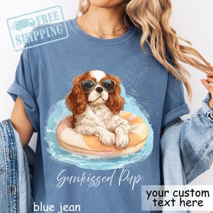 Pode incluir: Camiseta azul jeans com um desenho de um Cavalier King Charles Spaniel usando óculos de sol e flutuando em uma piscina em uma boia laranja e branca. O texto "Sunkissed Pup" está abaixo. O texto "your custom text here" também é visível.