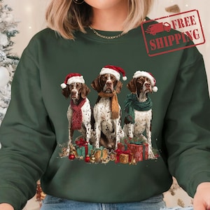 May include: Green sweatshirt featuring three dogs wearing Santa hats and scarves. The dogs are German Shorthaired Pointers with brown and white markings. Presents and ornaments are at the bottom of the image. The words "FREE SHIPPING" are in a red stamp.
