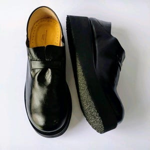May include: A pair of black leather platform shoes with a thick rubber sole. The shoes have a simple design with a rounded toe and a strap across the top.
