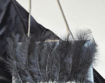 May include: A rectangular clutch with a silver frame and a delicate chain strap. The clutch is adorned with black feathers, creating a textured and elegant appearance. The background is a dark, velvety fabric.