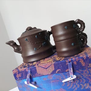 Peut inclure: Une théière en céramique brune et deux tasses assorties avec des motifs floraux, le tout dans une boîte à motifs violets et dorés.