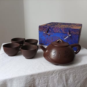 Peut inclure: Un service à thé en céramique marron comprenant une théière, quatre tasses et une boîte décorative. La théière a une anse et un bec courbés, avec des caractères chinois gravés sur le côté. La boîte est bleue avec un motif floral.