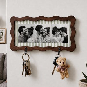 May include: A rectangular wall-mounted key holder with a dark brown wavy wooden frame. The center features three black and white photographs of a couple against a light green and white striped background. Two black metal hooks hold keys.