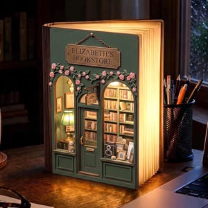 May include: A green book-shaped diorama lamp titled Elizabeth's Bookstore, featuring a miniature storefront with pink floral vines, an arched door, and interior shelves filled with books, illuminated by warm light from within the pages.