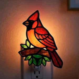 May include: A stained glass night light shaped like a cardinal bird perched on a branch. The bird features vibrant red and orange glass panels, while the branch is accented with green leaves. The light emits a warm glow from the wall outlet.