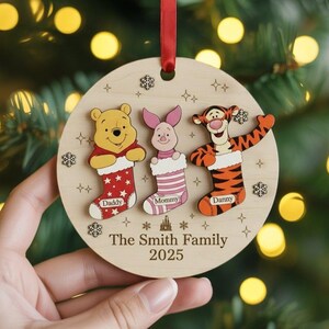 May include: A wooden Christmas ornament featuring Winnie the Pooh, Piglet, and Tigger characters. Each character is above a stocking with a name. The ornament has a red ribbon for hanging and reads "The Smith Family 2025".