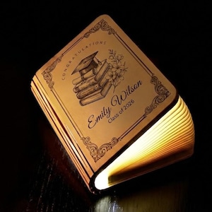 Può includere: Una lampada a forma di libro in legno che emette una luce calda. La copertina è incisa con un cappello di laurea, libri, fiori e il testo "Congratulations Emily Wilson Class of 2026". La lampada è aperta, rivelando le pagine illuminate.