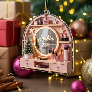 May include: A pink vanity Christmas ornament with a mirror, miniature makeup items, and a small Christmas tree. The mirror is inscribed with the name "Sophia" and the year "2025". The ornament is hanging from a gold string.