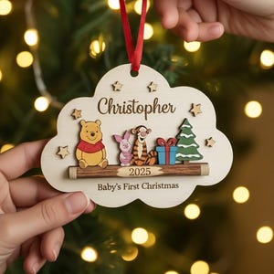 May include: A wooden Christmas ornament in the shape of a cloud, featuring Winnie the Pooh, Piglet, Tigger, a Christmas tree, and a gift. The ornament has the name "Christopher" and the text "Baby's First Christmas" with the year 2025.