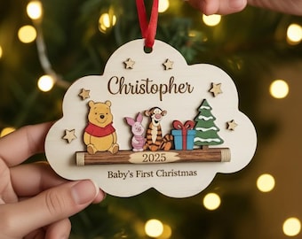 May include: A cloud-shaped wooden Christmas ornament featuring Winnie the Pooh, Piglet, Tigger, a gift, and a tree. The text reads Christopher, 2025, and Baby's First Christmas. It hangs from a red ribbon against a background of blurred golden lights.