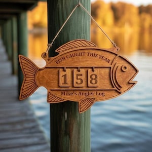 Puede incluir: Letrero de madera con forma de pez con el texto "FISH CAUGHT THIS YEAR" y "Mike's Angler Log". El letrero muestra el número "158". Está colgado de un poste de madera con un fondo borroso de lago.