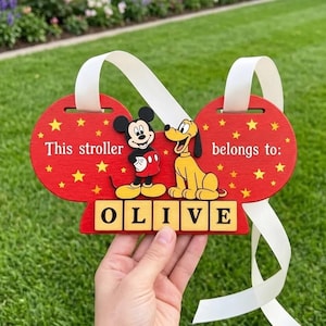 May include: A red wooden stroller sign shaped like mouse ears featuring Mickey Mouse and Pluto. The text reads "This stroller belongs to:" with the name "OLIVE" displayed on five individual square tiles. A white ribbon is attached for hanging.