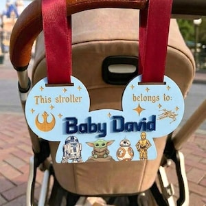 Peut inclure: Un porte-&eacute;tiquette de poussette bleu clair en forme d'oreilles de Mickey, avec le texte "This stroller belongs to: Baby David." Comprend des personnages et symboles de Star Wars. Attach&eacute; &agrave; une poign&eacute;e de poussette avec un ruban rouge.