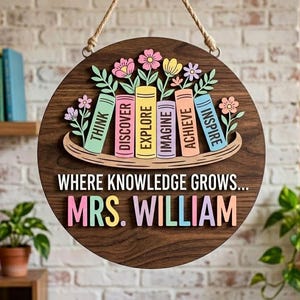 Könnte beinhalten: Runder Holzschild mit dunkelbraunem Finish, mit bunten Büchern und Blumen verziert. Das Schild zeigt den Text "WHERE KNOWLEDGE GROWS... MRS. WILLIAM" in Pastellfarben. Ein Seil ist zum Aufhängen befestigt.