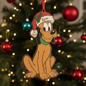 Puede incluir: Un adorno plano de madera para árbol de Navidad con la forma del perro de dibujos animados Pluto, que lleva un gorro de Papá Noel rojo con una ramita de acebo verde y el nombre Lucas impreso en el borde. Una pequeña etiqueta muestra el año 2025.