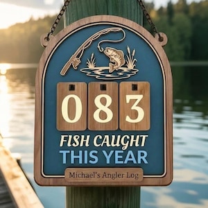 Puede incluir: Letrero de madera con temática de pesca con fondo azul, que presenta una caña de pescar y un pez saltando fuera del agua. El letrero muestra el número "083" y el texto "FISH CAUGHT THIS YEAR". Incluye el texto "Michael's Angler Log".