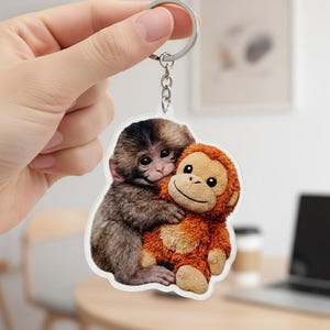 May include: A keyring featuring a cute monkey hugging a stuffed monkey. The keyring has a silver ring and chain. The monkeys are brown and orange, with a smiling face on the stuffed monkey.