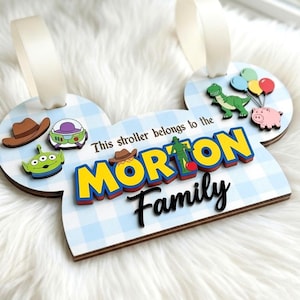 Könnte beinhalten: Ein hellblau-weiß kariertes Kinderwagenschild in Form von Mausohren mit zwei weißen Bandschlaufen. Es zeigt Comic-Figuren und den Text "This stroller belongs to the MORTON Family" in fetter gelber, roter und schwarzer Schrift.