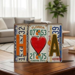 May include: Decorative license plate letters spelling "H-E-A". The "H" is from Texas, the "E" is a red heart from Florida, and the "A" is from California. Each letter is a different color and design, creating a unique home decor item.