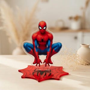 May include: A clear acrylic Spider-Man figure in a crouching pose, with red and blue costume details. The figure is mounted on a red spiderweb base with a black spider emblem. The background is blurred.