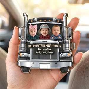 Peut inclure: Un ornement en forme de camion noir avec trois visages d'enfants dans le pare-brise. Le texte indique : KEEP ON TRUCKING, DADDY, We Love You, Noah, Chloe, James. Le design comprend des pots d'échappement argentés, des phares et une calandre.