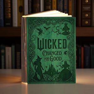 May include: A green wooden book-shaped lamp with the text "Wicked Changed for Good" on the cover. The cover features illustrations of characters and symbols from the musical. The pages are illuminated, creating a warm glow.