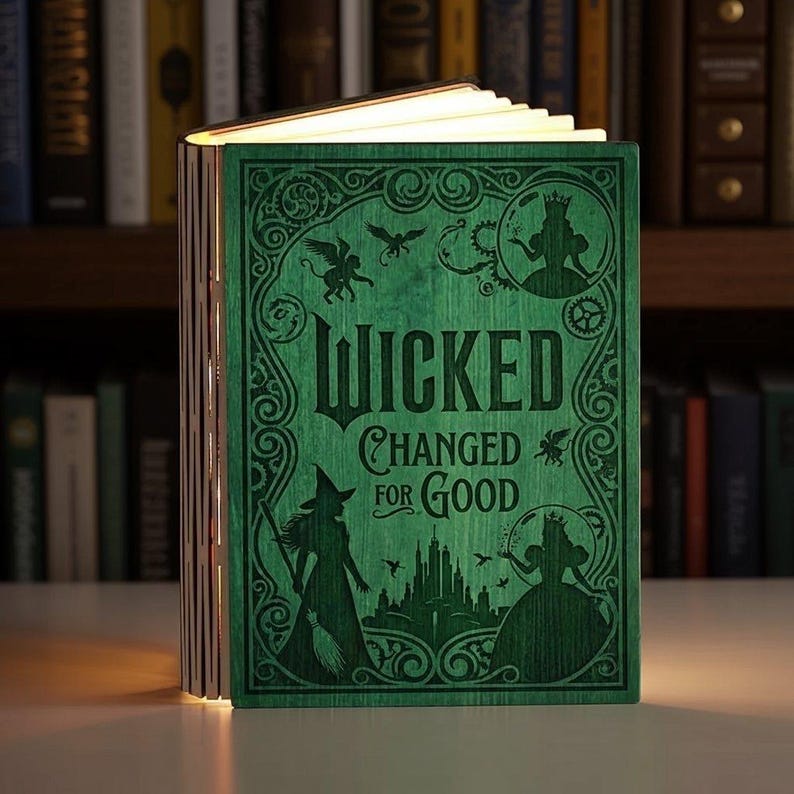 May include: A green wooden book-shaped lamp with the text "Wicked Changed for Good" on the cover. The cover features illustrations of characters and symbols from the musical. The pages are illuminated, creating a warm glow.