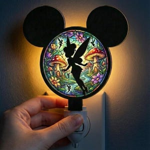 May include: A decorative night light featuring a silhouette of Tinkerbell in a colorful fairy garden scene. The night light has a black Mickey Mouse head shape and plugs directly into a wall outlet.
