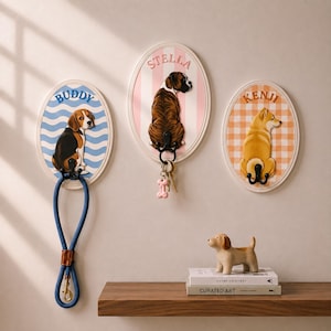 Könnte beinhalten: Drei ovale Wandhaken mit Hundeillustrationen. Die linke Tafel zeigt einen Beagle mit der Aufschrift BUDDY auf blauem Wellenhintergrund. Die mittlere Tafel zeigt einen Boxer mit der Aufschrift STELLA auf rosa Streifen. Die rechte Tafel zeigt einen Hund mit der Aufschrift KENJI auf orangefarbenem Karomuster. Jede Tafel hat einen schwarzen Metallhaken.