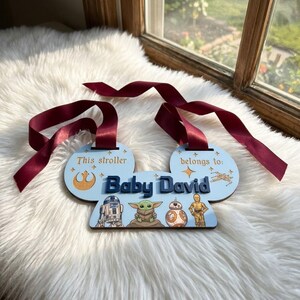 Peut inclure: Une &eacute;tiquette de poussette bleu clair avec le texte "This stroller belongs to: Baby David." Repr&eacute;sente les personnages de Star Wars R2-D2, Grogu, BB-8 et C-3PO. Comprend un ruban bordeaux pour l'accrocher.