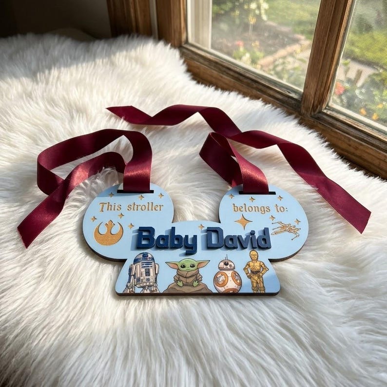 Peut inclure: Une &eacute;tiquette de poussette bleu clair avec le texte "This stroller belongs to: Baby David." Repr&eacute;sente les personnages de Star Wars R2-D2, Grogu, BB-8 et C-3PO. Comprend un ruban bordeaux pour l'accrocher.