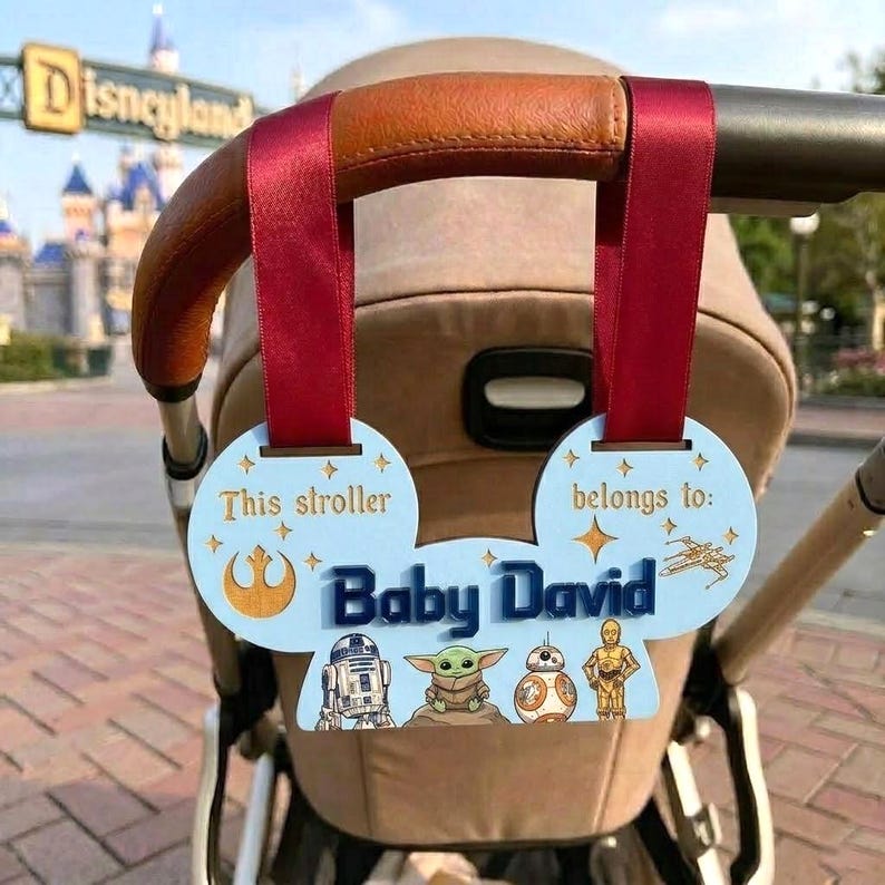 Peut inclure: Un porte-&eacute;tiquette de poussette bleu clair en forme d'oreilles de Mickey, avec le texte "This stroller belongs to: Baby David." Comprend des personnages et symboles de Star Wars. Attach&eacute; &agrave; une poign&eacute;e de poussette avec un ruban rouge.