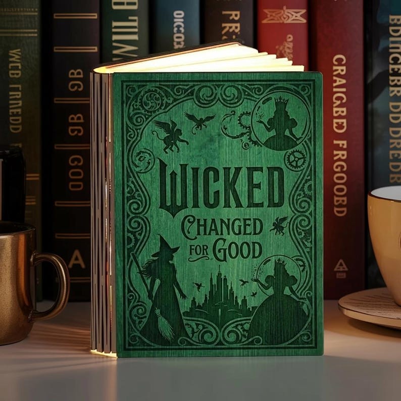May include: A green wooden book-shaped lamp with the words "WICKED CHANGED FOR GOOD" engraved on the cover. The cover features silhouettes of characters and decorative elements. The lamp is open, emitting a warm glow, and is surrounded by other books.