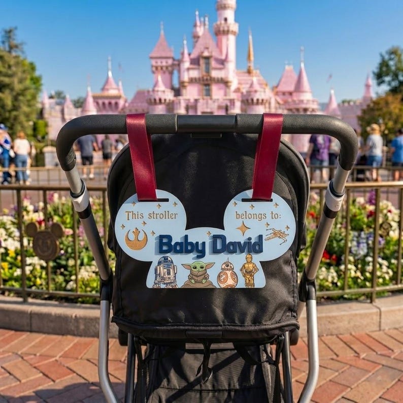 Peut inclure: Une poussette noire avec une &eacute;tiquette personnalis&eacute;e sur le th&egrave;me de Star Wars. L'&eacute;tiquette est bleu clair avec le texte "Baby David" et des personnages de Star Wars. La poussette a des sangles bordeaux.