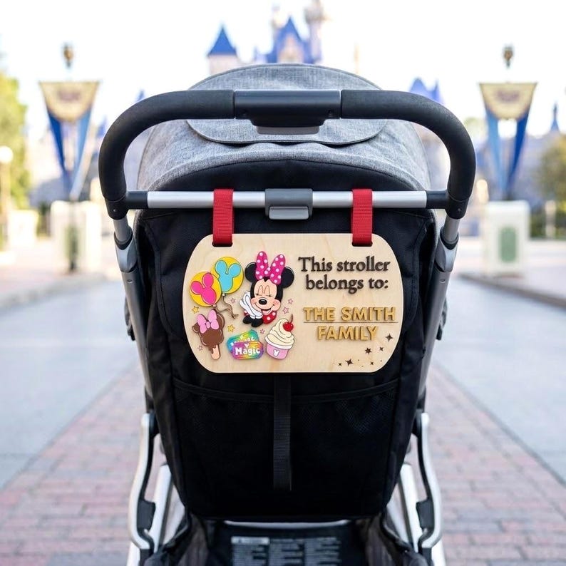 May include: A light-colored wooden stroller sign attached with red straps to a black stroller. The sign features a Minnie Mouse graphic, colorful balloons, a cupcake, and the text: This stroller belongs to: THE SMITH FAMILY in black lettering.