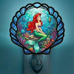 Puede incluir: Luz de noche decorativa con un diseño de vidriera de una sirena con cabello rojo y cola verde, sentada en una roca. La sirena está dentro de un marco de concha azul, con peces y coral en el fondo.