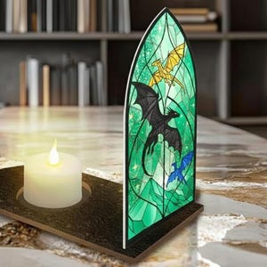 May include: A decorative stained glass panel with a dragon design in green, black, gold, and blue. A white candle sits in a dark brown holder. The panel is shaped like a church window.