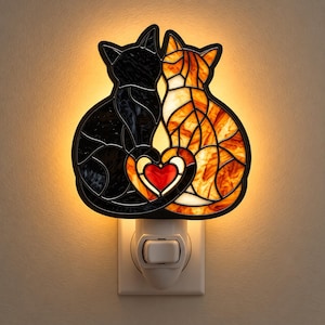 May include: A stained glass night light featuring two cats sitting side by side, one black and one orange tabby, with their tails forming a heart shape around a central red heart. The lamp emits a warm glow from behind the glass panels.